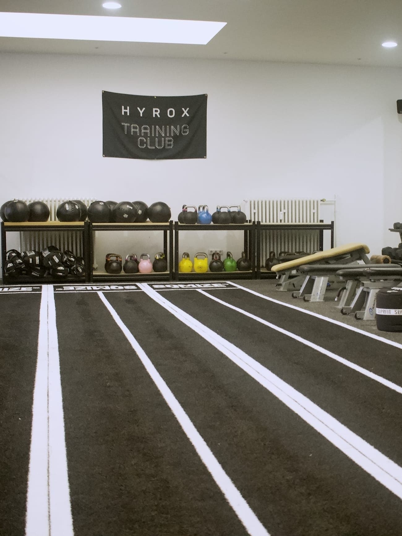 Die HYROX-Wand des Studios mit Wall Balls und Kettlebells im Regal, davor die markierten Bahnen